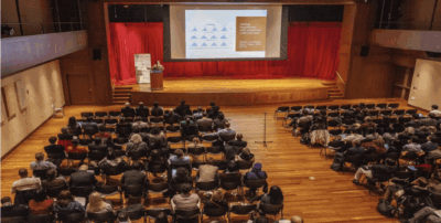 A large audience sits in an auditorium facing a stage with red curtains, where a person is giving a presentation in front of a screen displaying slides. The photo is viewed from the back of the room.