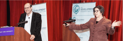 Two people stand at separate podiums on a stage, speaking into microphones. Both are smiling. Behind them are banners with the text Development Research Institute. A red curtain hangs in the background.