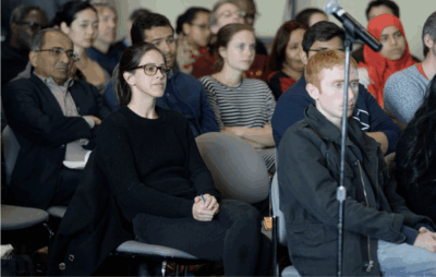 A diverse group of people sits attentively in rows of chairs at an indoor event, with a microphone stand visible in the foreground. The attendees appear focused on a speaker or presentation.