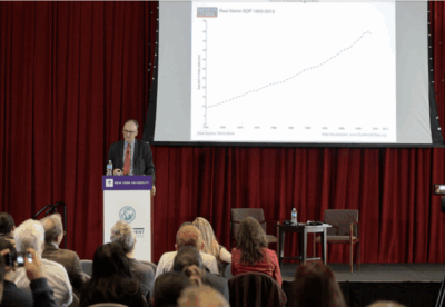 A speaker stands at a podium presenting to an audience in front of red curtains, with a line graph displayed on a screen behind them. Several audience members are taking photos or videos.