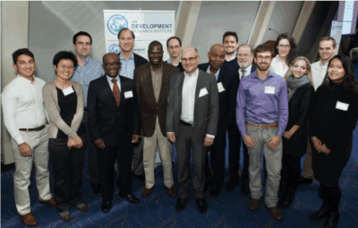 A group of sixteen people, both men and women, are standing together indoors, smiling for a group photo at a professional event. A sign in the background reads Development.