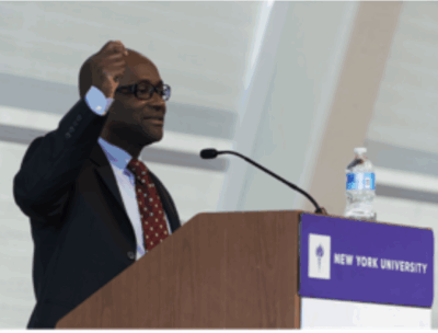 A man in a suit and glasses speaks at a podium with a New York University sign, gesturing with one hand, in a modern indoor setting.