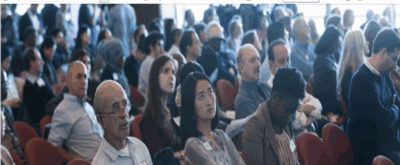 A large group of people sits in rows of red chairs at an indoor event, attentively listening. Some are looking forward, some are taking notes, and the audience is diverse in age, gender, and ethnicity.