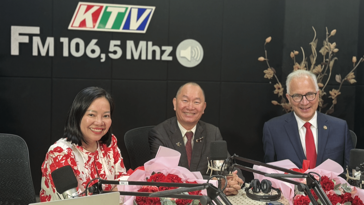 Three people in formal attire sit at a radio station table with microphones and flower bouquets. The black wall behind them displays the logo KTV FM 106,5 Mhz and decorative branches. All three are smiling at the camera.
