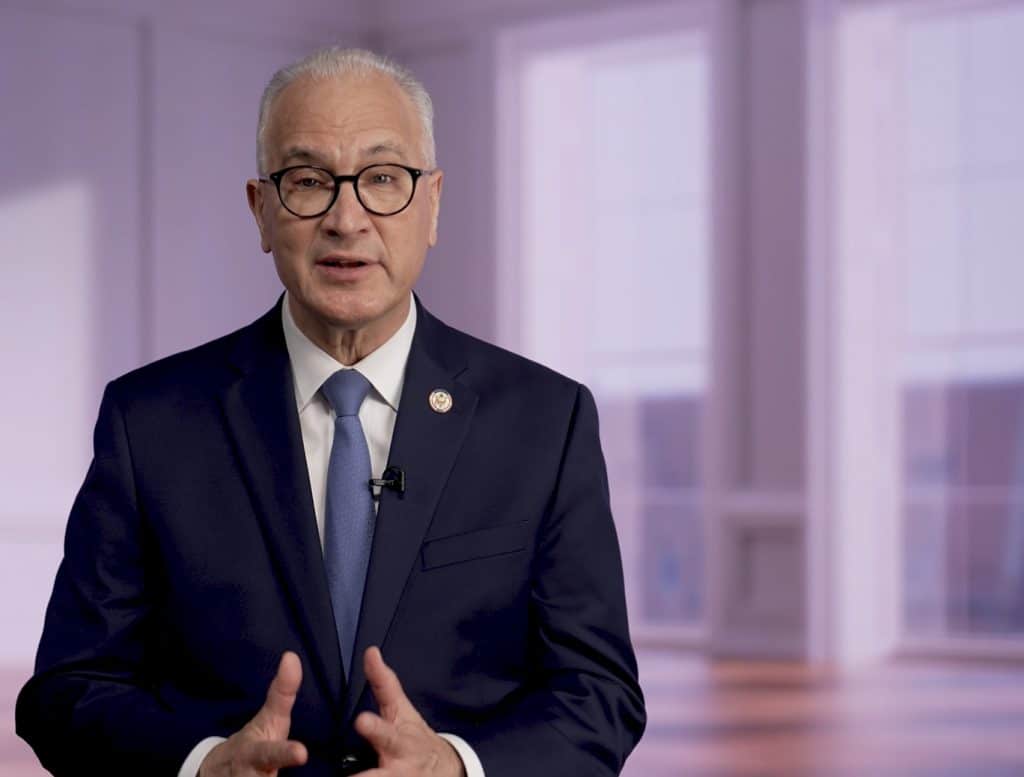 An older man in a dark suit, white shirt, and blue tie is speaking and gesturing with his hands about the WISC. He wears glasses and stands in a brightly lit room with large windows and a blurred background.