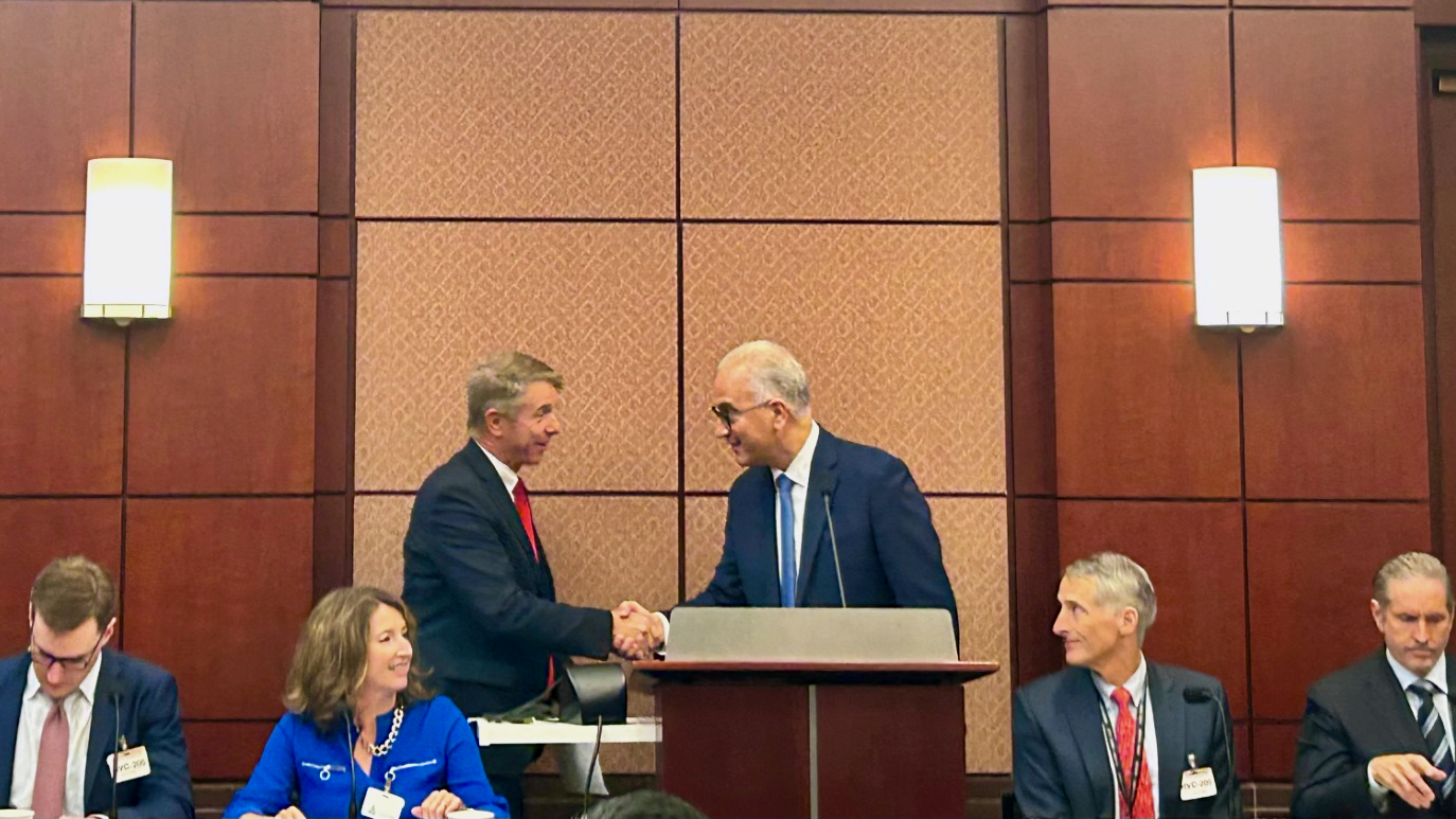 Six people in business attire are seated or standing at a conference table. Two men at the center are smiling and shaking hands behind a podium, while others look on and interact. The setting appears formal, with wood-paneled walls and lights.